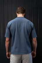 Man met een blauw T-shirt en een grijze broek tegen een zwarte achtergrond.