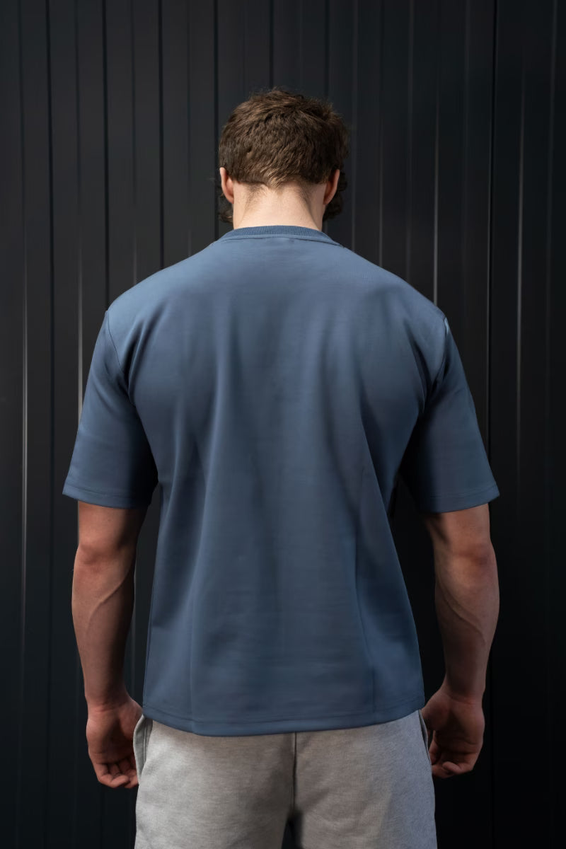 Man met een blauw T-shirt en een grijze broek tegen een zwarte achtergrond.