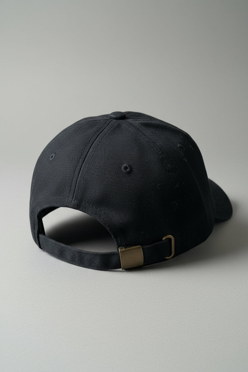 Rear three-quarter view of the OMW (on my weights) black dad hat resting on a light-gray studio floor, showing the adjustable strap with brass buckle and D-ring, subtle dust specks on pitch-black cotton twill fabric, ultra-sharp product photography.