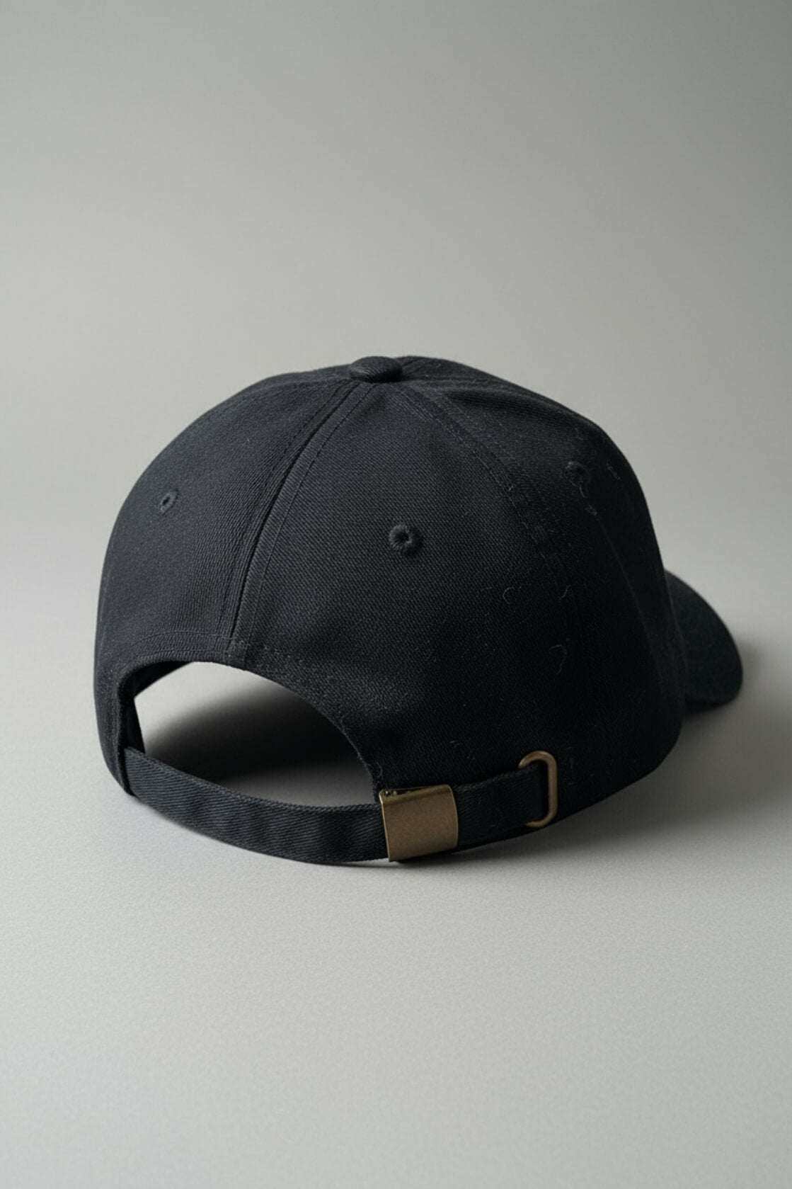 Rear three-quarter view of the OMW (on my weights) black dad hat resting on a light-gray studio floor, showing the adjustable strap with brass buckle and D-ring, subtle dust specks on pitch-black cotton twill fabric, ultra-sharp product photography.