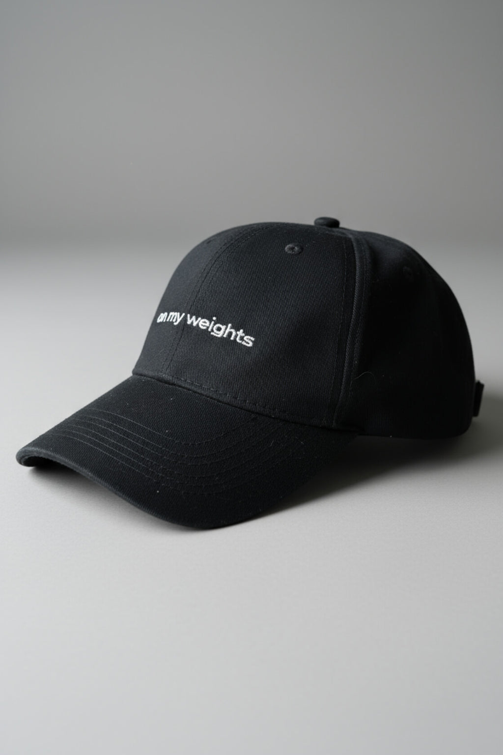 Right-side three-quarter view of the OMW black dad hat laid flat on a neutral light-gray studio floor, white “on my weights” script partially visible, realistic fabric texture with micro-lint and dust, ultra-sharp 3D render.