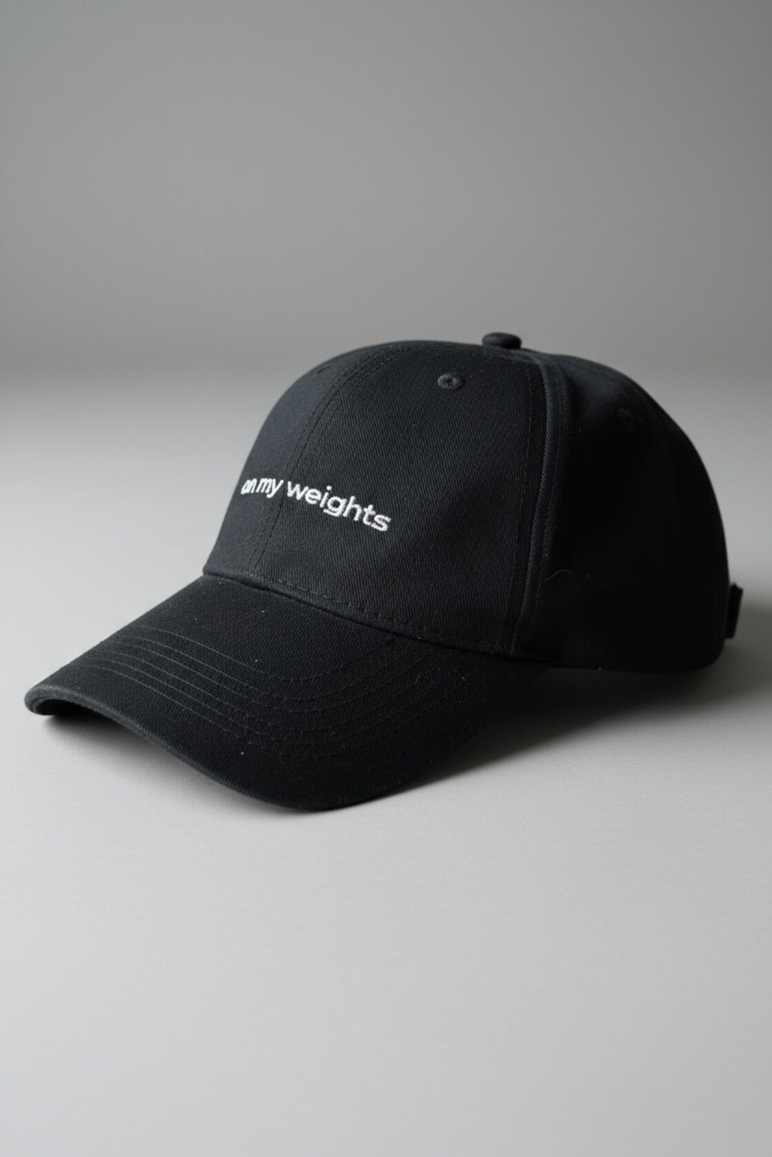 Right-side three-quarter view of the OMW black dad hat laid flat on a neutral light-gray studio floor, white “on my weights” script partially visible, realistic fabric texture with micro-lint and dust, ultra-sharp 3D render.