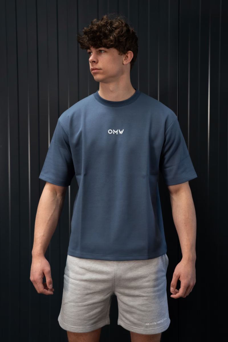Persoon met een blauw T-shirt met logo en een grijze korte broek tegen een zwarte achtergrond.