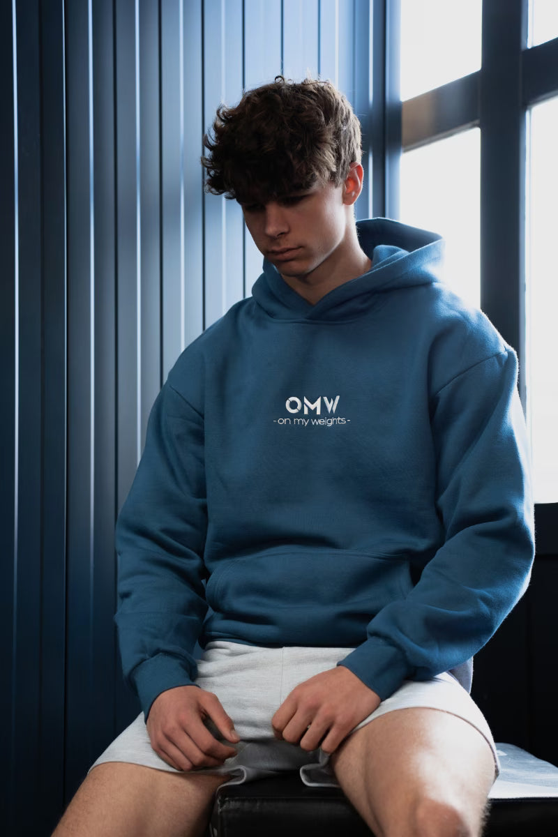 Een persoon met een blauwe hoodie met het 'OMW'-logo zit tegen een muur met een raam.