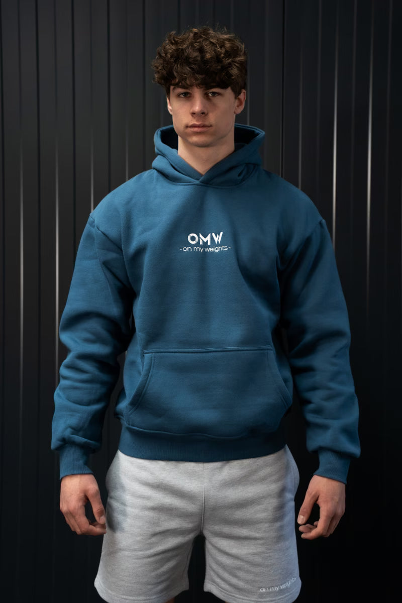 Persoon met een blauwe hoodie met 'OMW'-logo geborduurd, staandvoor een donkere achtergrond.
