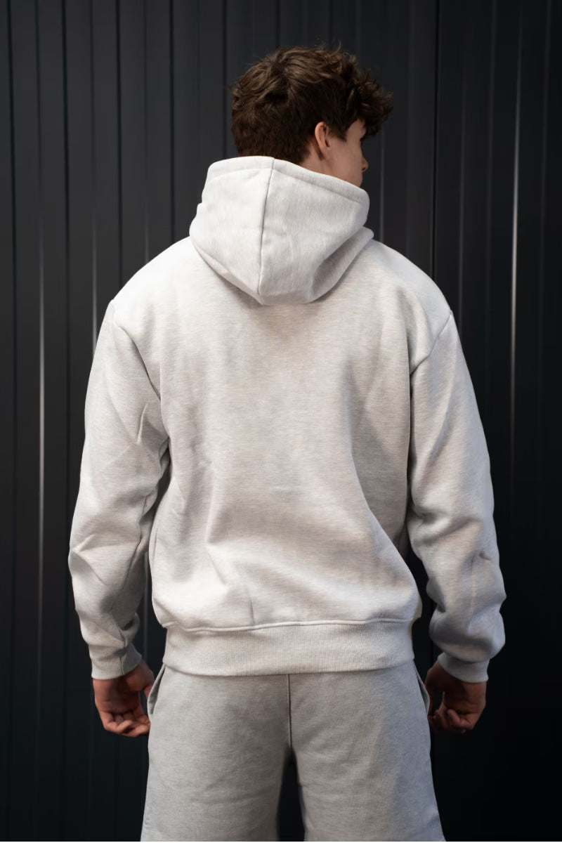 Persoon met een grijze hoodie en broek voor een donkere achtergrond.