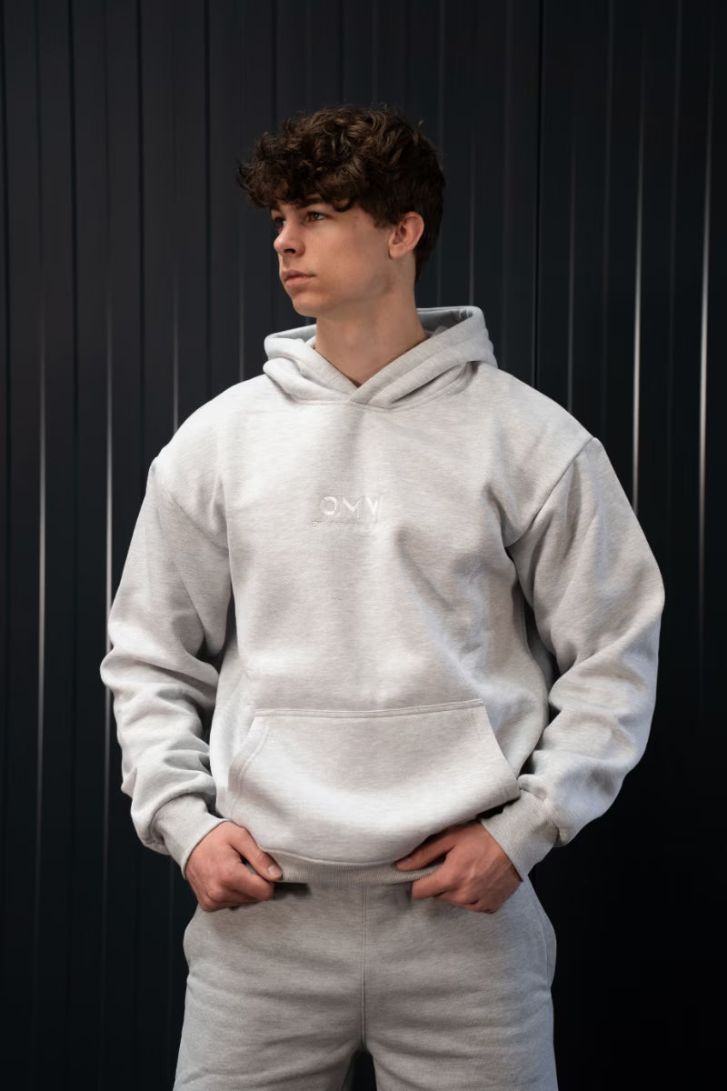 Persoon met een grijze hoodie en broek voor een zwarte achtergrond.