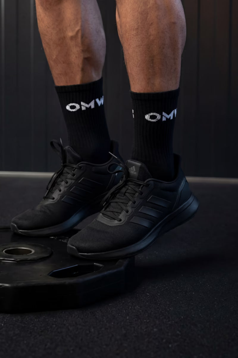 Persoon met zwarte sneakers en sokken met zichtbaar logo op een donkere achtergrond.