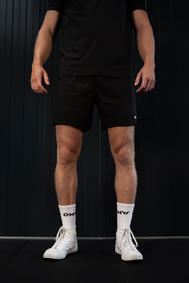 Persoon met zwarte sportbroek, witte sokken met logo en witte sneakers tegen een zwarte achtergrond.