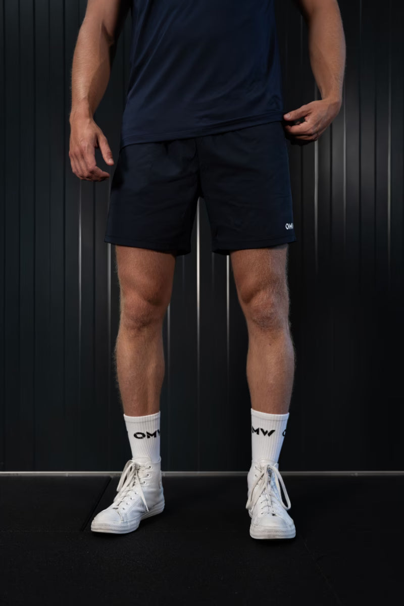 Persoon met donkerblauwe korte broek en witte sneakers met zichtbaar logo, tegen een zwarte achtergrond.