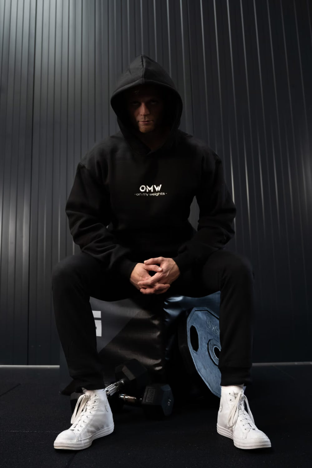 Een persoon met een zwarte hoodie met het 'OMW'-logo zit op een blauwe gewichtsschijf tegen een donkere achtergrond.