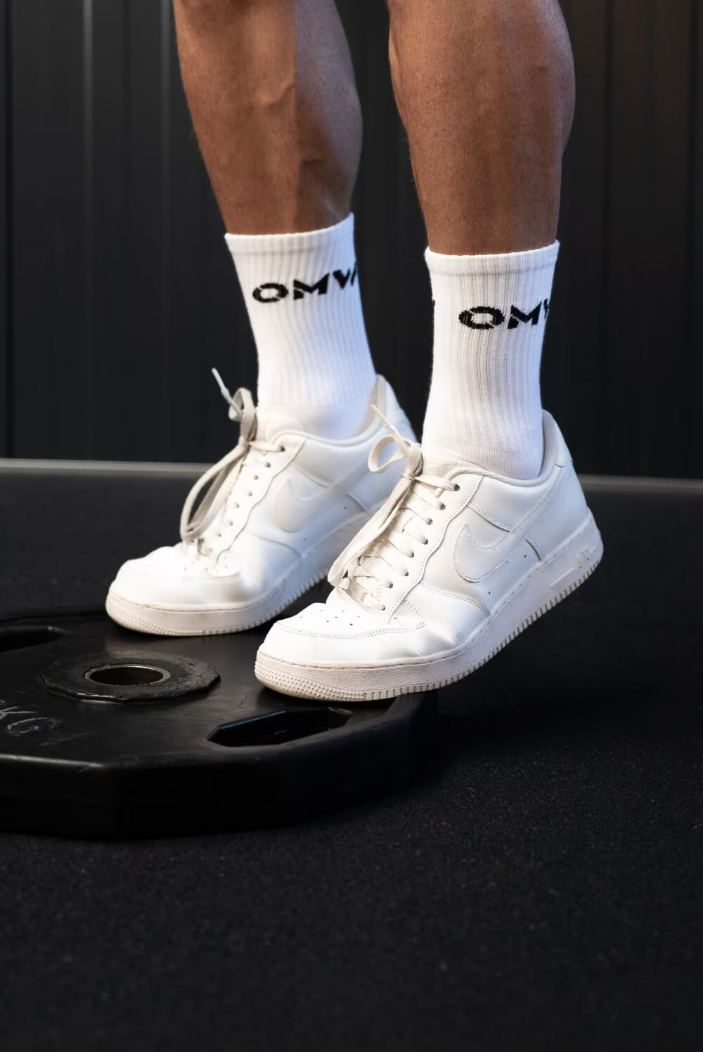 Witte sneakers en sokken met 'OMW' logo voor een zwarte achtergrond, staand op een gewicht.