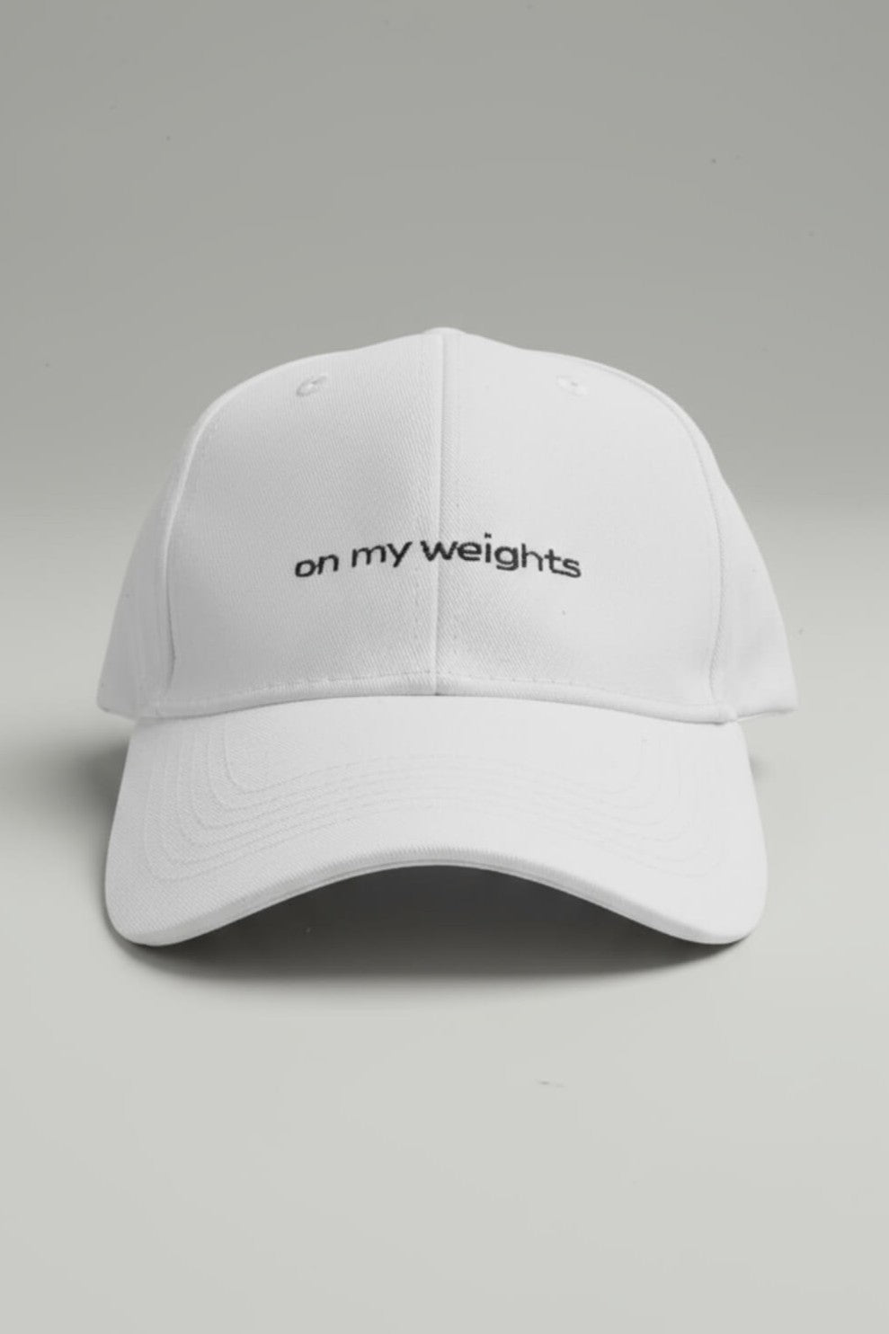 Front view of the OMW white dad hat centered on a light-gray seamless surface, white “on my weights” embroidery prominently displayed on the front panel, bill curved naturally, soft drop shadow, high-detail product shot.