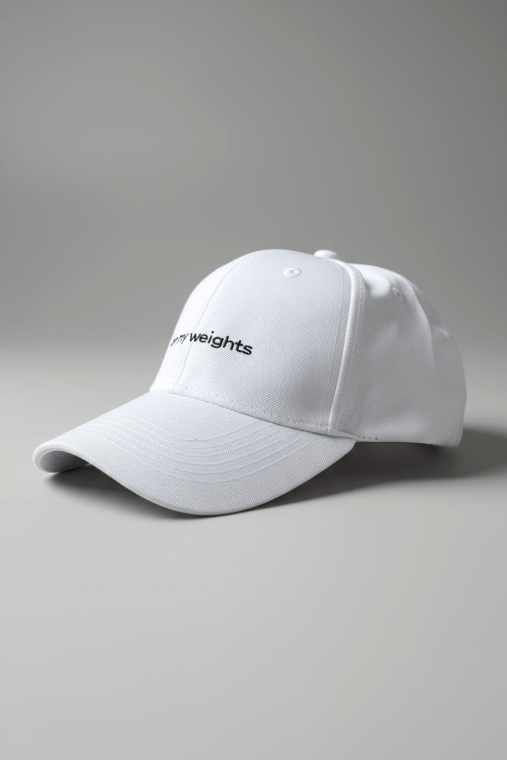 Right-side three-quarter view of the OMW white dad hat laid flat on a neutral light-gray studio floor, white “on my weights” script partially visible, realistic fabric texture with micro-lint and dust, ultra-sharp 3D render.