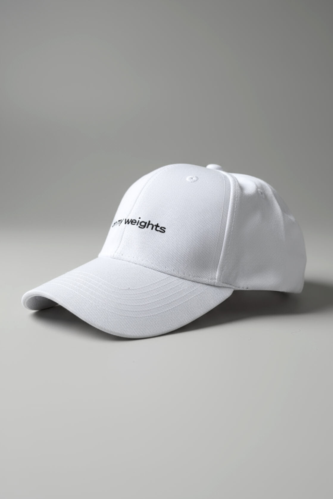 Right-side three-quarter view of the OMW white dad hat laid flat on a neutral light-gray studio floor, white “on my weights” script partially visible, realistic fabric texture with micro-lint and dust, ultra-sharp 3D render.