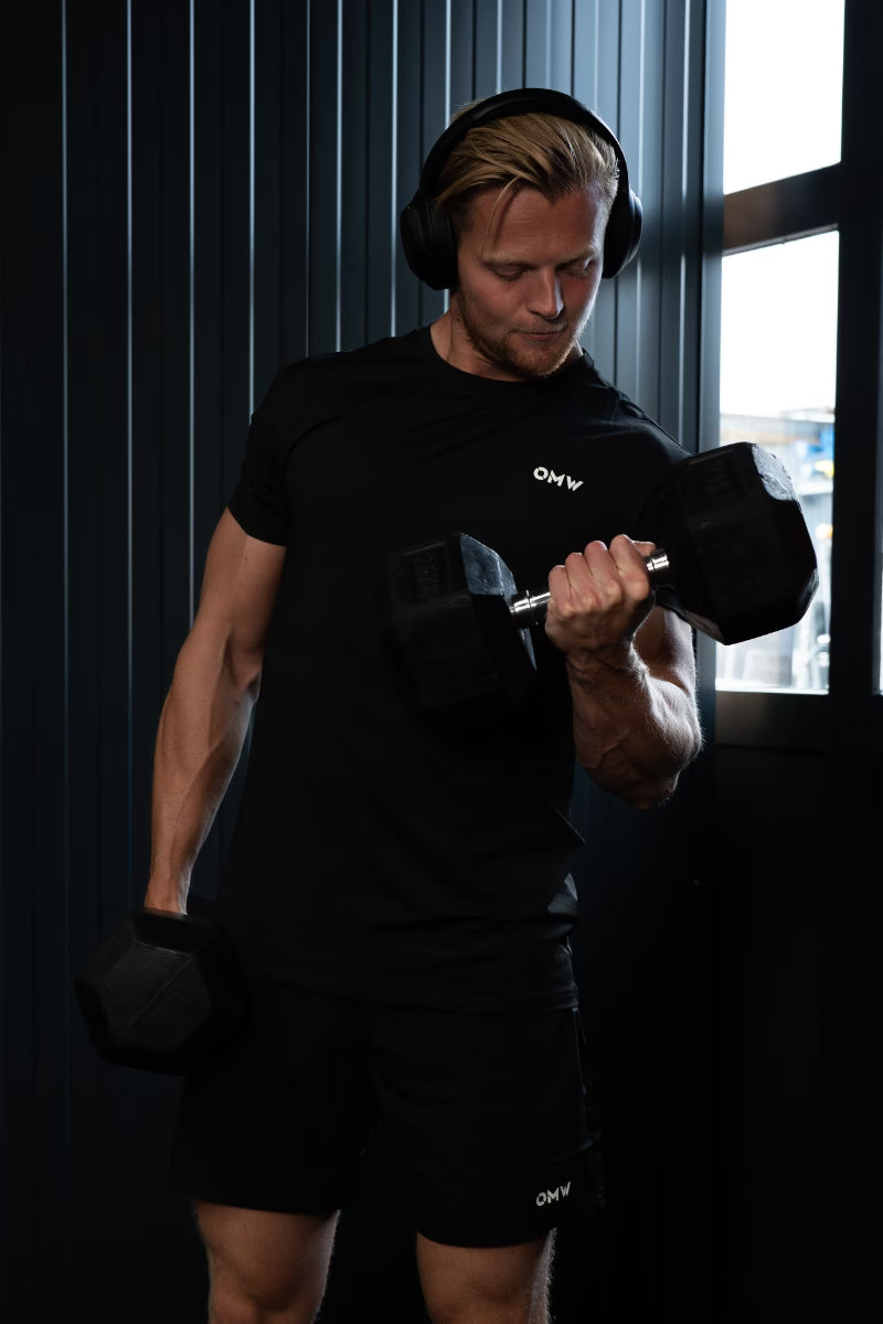 Een man, gekleed in een zwart sport T-shirt en een korte broek met een zichtbaar merklogo 'OMW', is aan het trainen met dumbbells.