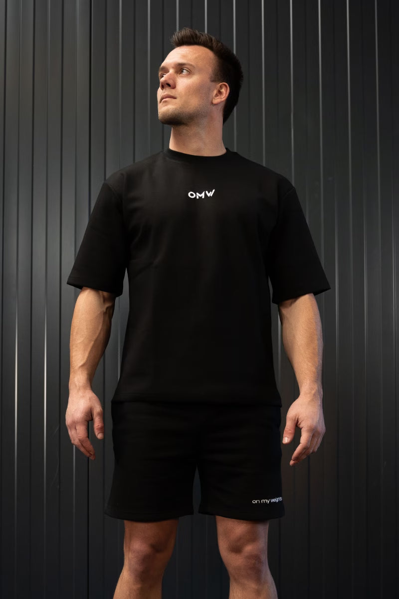 Man in een zwart t-shirt en korte broek met een OMW logo voor een donkere achtergrond.