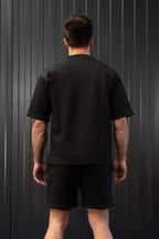 Een persoon in een zwart t-shirt en korte broek staat tegen een donkere muur met verticale strepen.