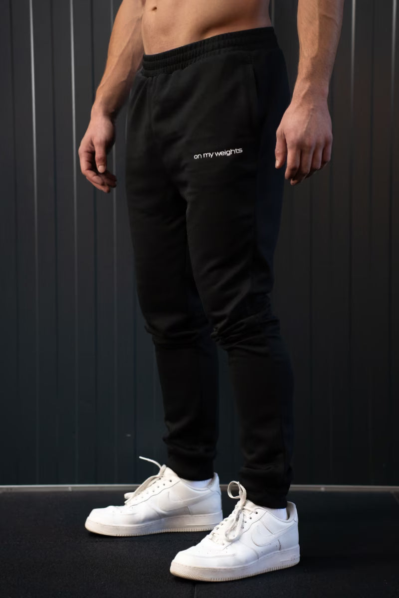 Persoon in zwarte joggingbroek met merklogo en witte sneakers tegen een donkere achtergrond.