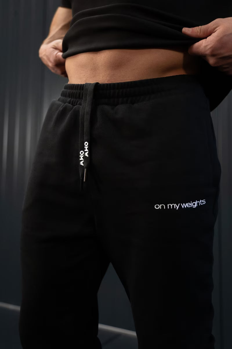 Persoon in zwarte joggingbroek met de tekst 'on my weights' op een donkere achtergrond.