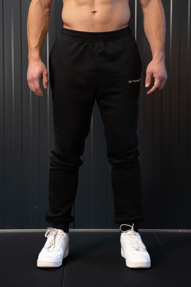 Persoon in zwarte joggingbroek met merklogo op een donkere achtergrond