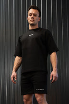 Man in een zwart katoenen t-shirt en korte broek met een geborduurd merklogo op een gestreepte achtergrond.