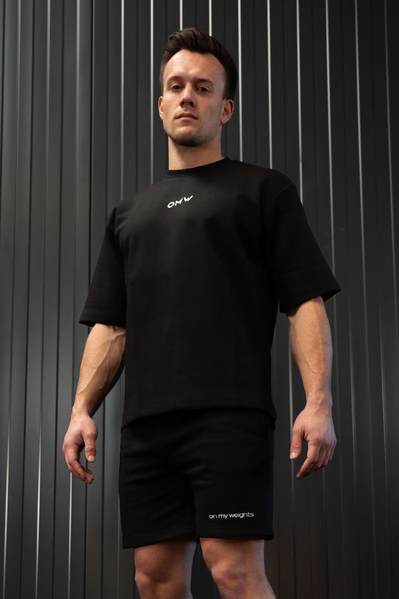 Man in een zwart katoenen t-shirt en korte broek met een geborduurd merklogo op een gestreepte achtergrond.