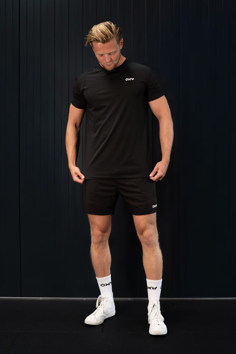 Man in zwarte sportkleding met zichtbare merklogo's van 'OMW' voor een zwarte achtergrond.