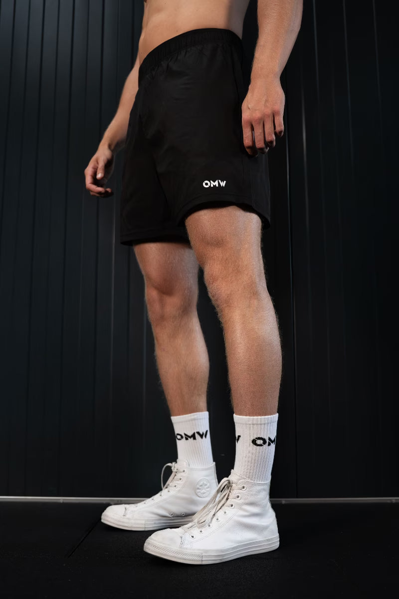 Persoon met zwarte korte broek met 'OMW'-logo, witte sokken en witte sneakers op een zwarte achtergrond.