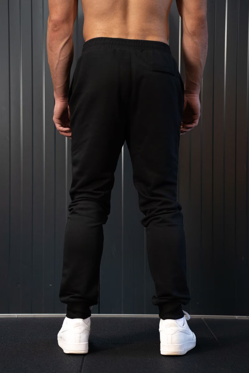 Persoon met zwarte joggingbroek en witte sneakers tegen een donkere achtergrond.